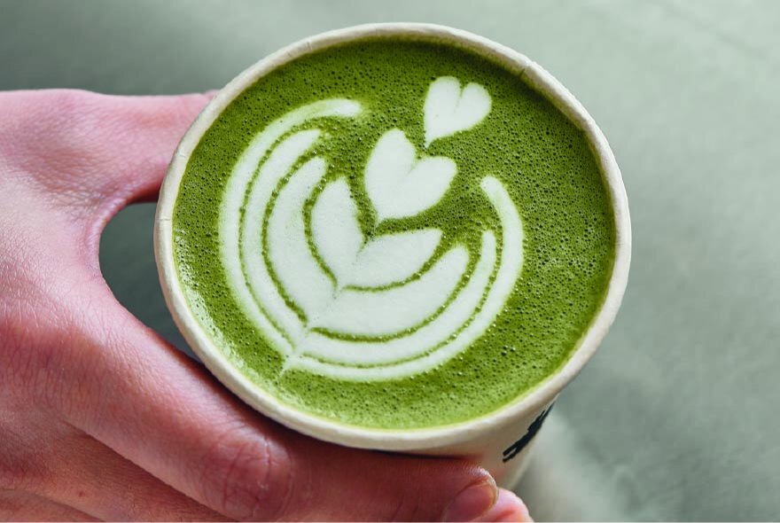 A hand holding a cup of green matcha latte with white foam art in a heart and leaf pattern.