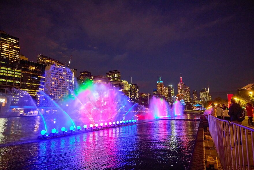 Coloured lights illuminate water sprays high in the air on the Yarra River at night.