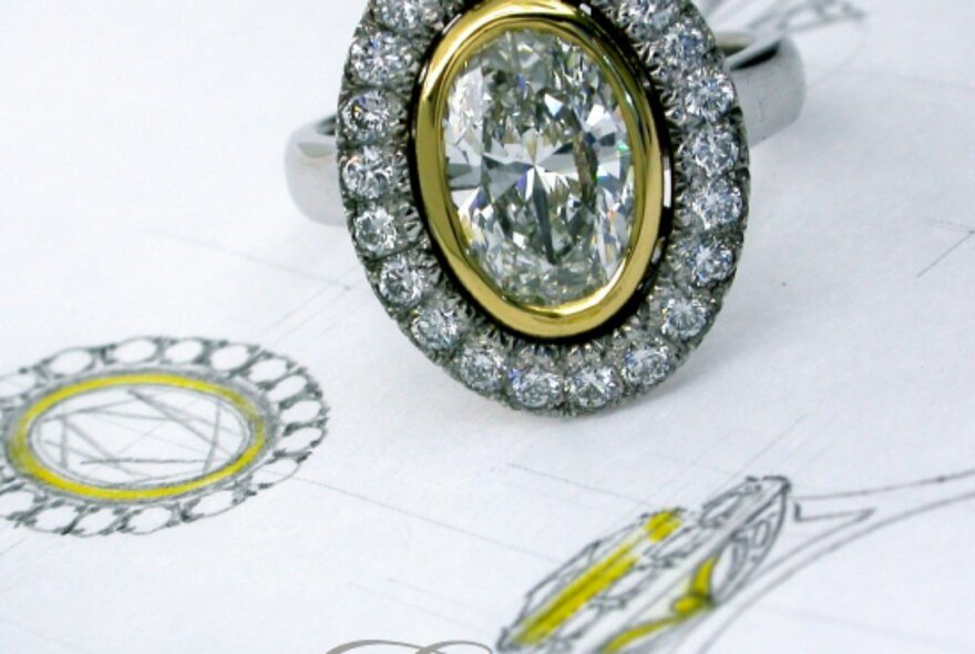 A diamond encrusted gold and white gold engagement ring with an oval cut diamond in the centre, resting on a paper with its matching hand-drawn design.