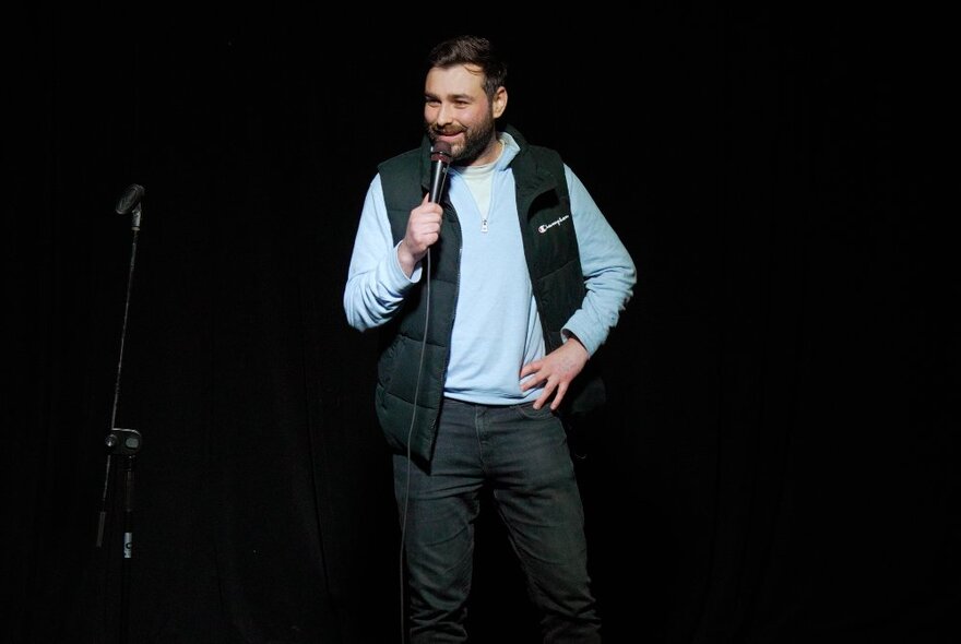 A man on stage with a microphone, all black surrounding him.