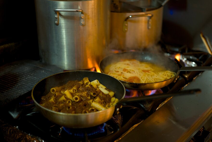 Two pans of pasta cooking on a gas burner in a commercial kitchen setting.
