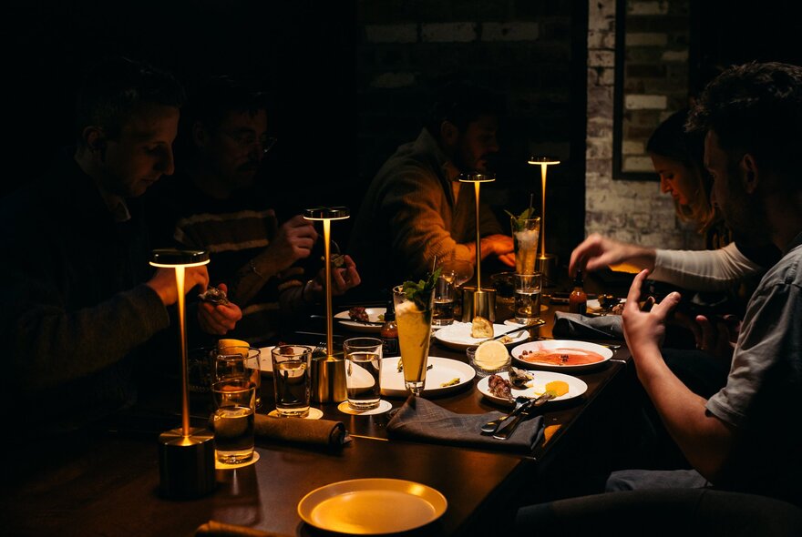 A dimly lit table at Death & Co with people enjoying drinks and shared plates of food, under the soft warm glow of individual table lamps.