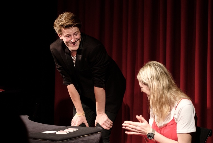 Magician Liam ‘LJ’ Jumpertz leaning forward on stage and performing a trick with a young blonde woman