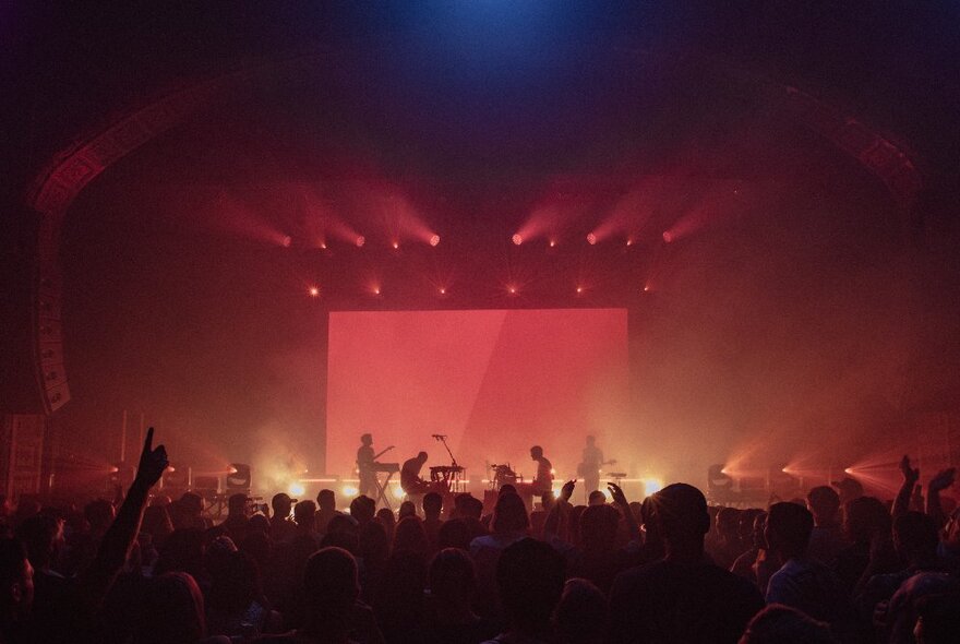 A smoky, red-lit stage with a band, seen from behind the silhouetted audience.