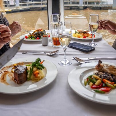 Diners seated at a restaurant table on a floating river cruise, with plates of  food in front of them and the water of the Yarra River in the background.