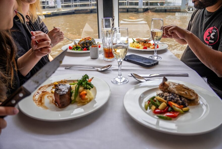 Diners seated at a restaurant table on a floating river cruise, with plates of  food in front of them and the water of the Yarra River in the background.
