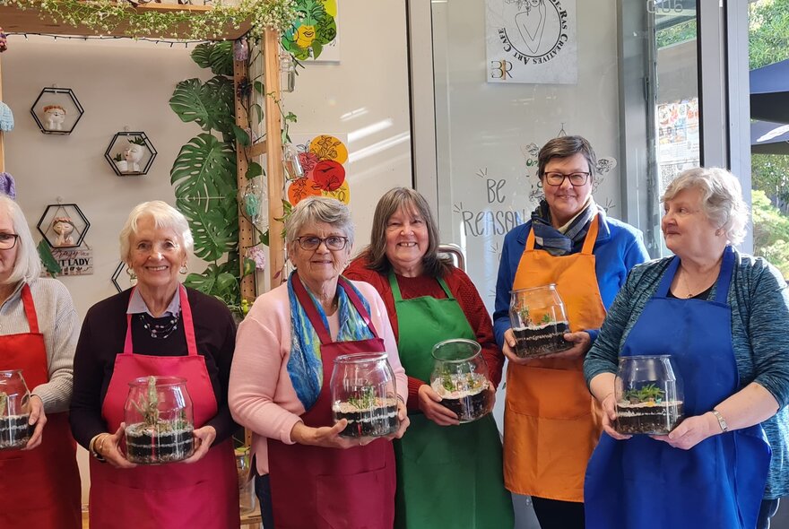 A group of smiling participants in a studio workshop setting, wearing colourful aprons and holding glass-domed terrariums.