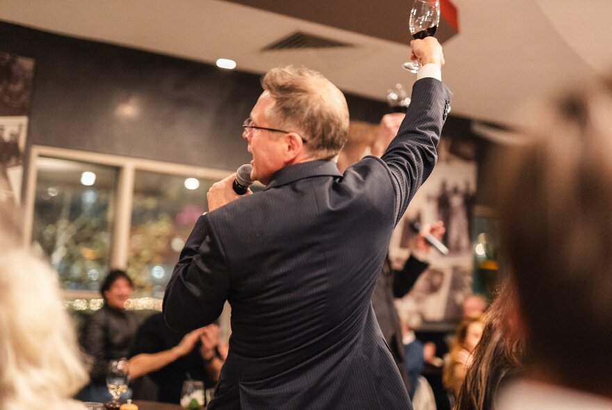 The back of a man in a suit holding a microphone in one hand and a glass of wine in the air. 