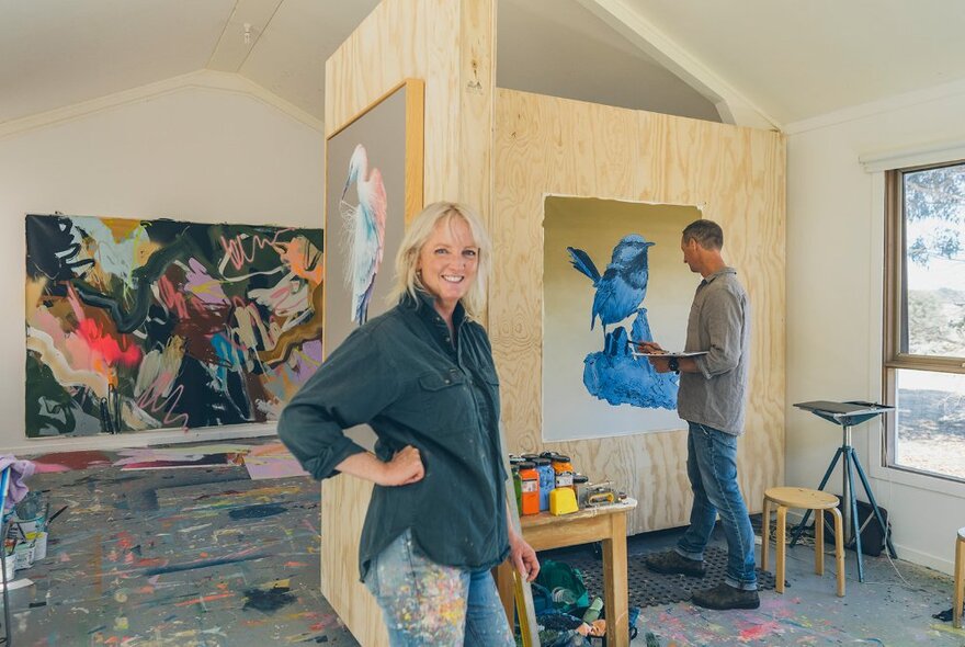 Artist standing with hand on hip in front of another artist painting a bluebird mural.