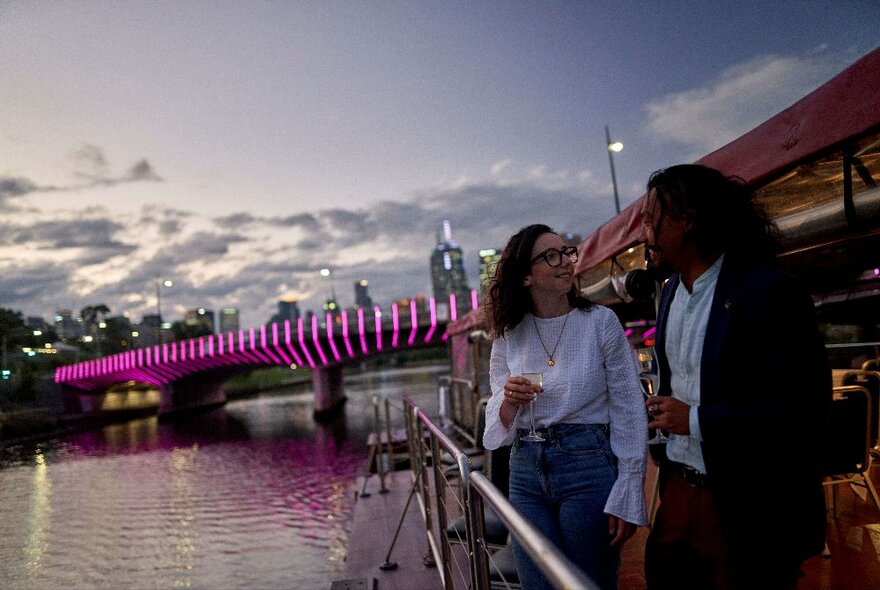 A couple standing on a boat deck looking at each other, holding a drink, with the river and an illuminated bridge in the background. 