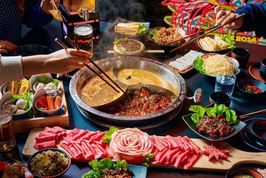 A pair of hands dipping food into a hot pot using chopsticks, with plates of raw meat and vegetables surrounding the hot pot.