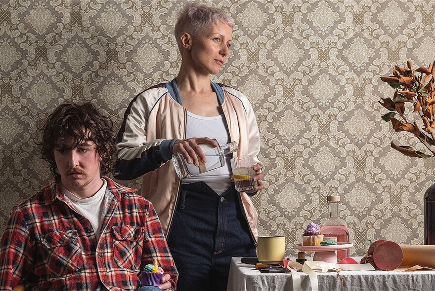 Actors in a scene of a play, person wearing plaid shirt seated at a table set with plates, person standing pouring water into a glass.