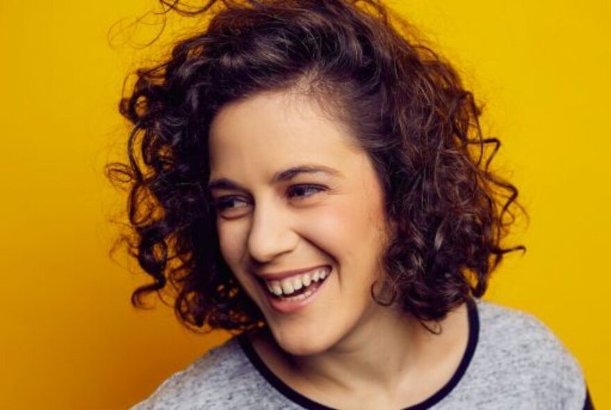 Laughing comedian with curly dark hair against a yellow background.