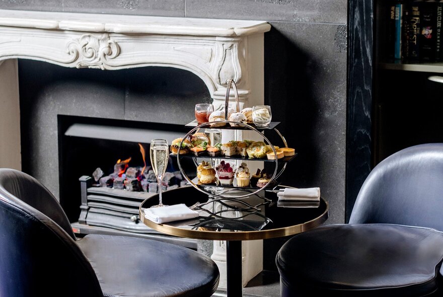 A high tea tray of treats on a two-seater table next to an open fireplace in a hotel with dark and moody styling.