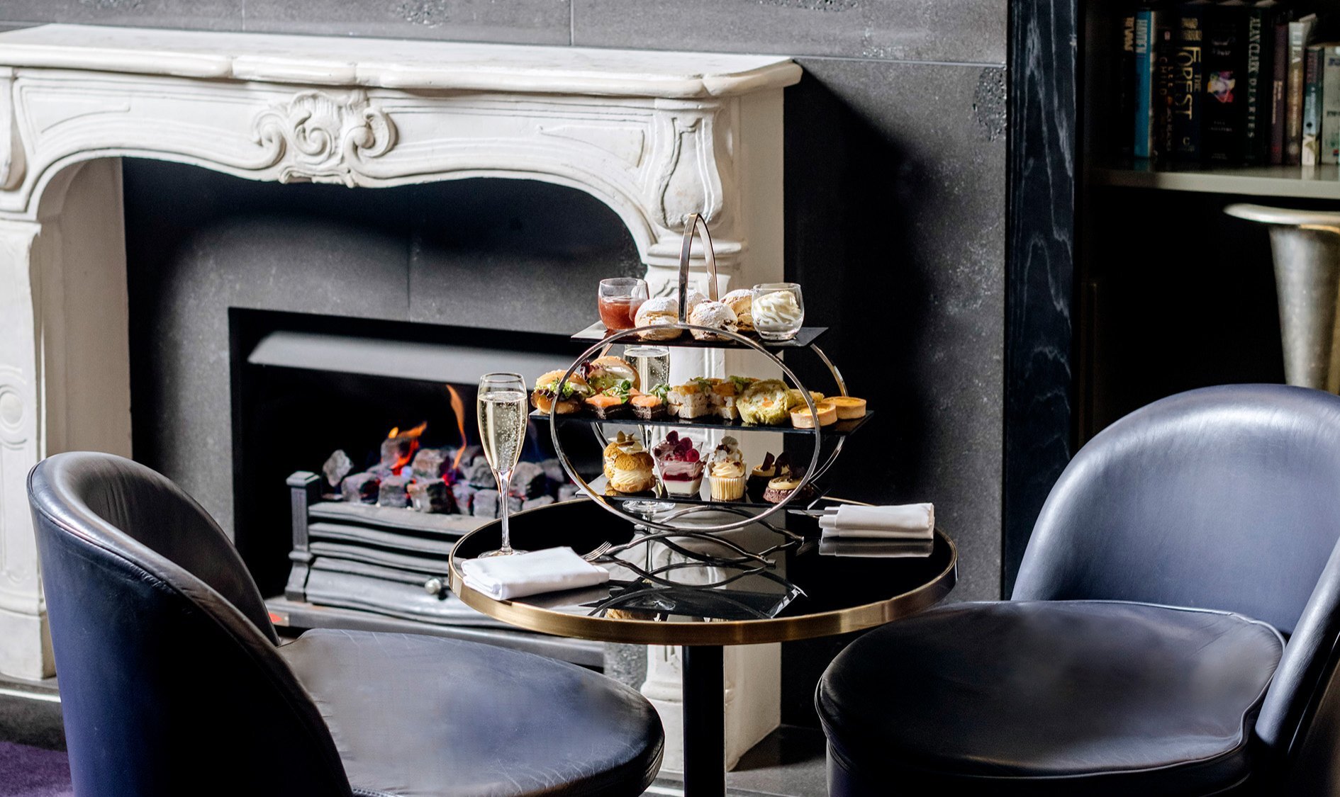 A high tea tray of treats on a two-seater table next to an open fireplace in a hotel with dark and moody styling.