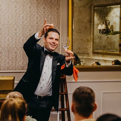 Man wearing a tuxedo performing sleight of hands tricks with a glass in front of a seated audience.