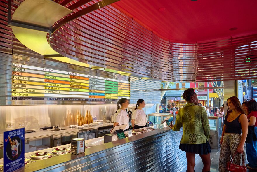 Two friends are inside a gelato shop. There are two servers waiting to take people's orders. There is a colourful menu of gelato flavours behind them. 