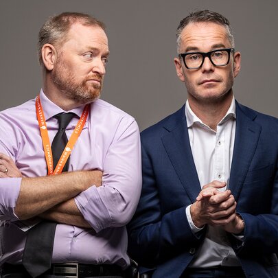 Two castmembers for Wankernomics sitting close together, one looking sideways at the other who is looking blankly ahead.