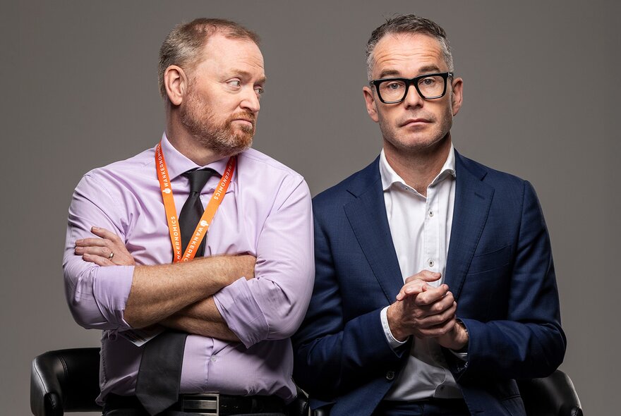 Two castmembers for Wankernomics sitting close together, one looking sideways at the other who is looking blankly ahead.