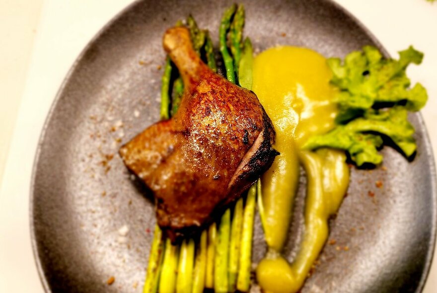 A plate with a joint of meat resting on top of grilled asparagus and green sauce.