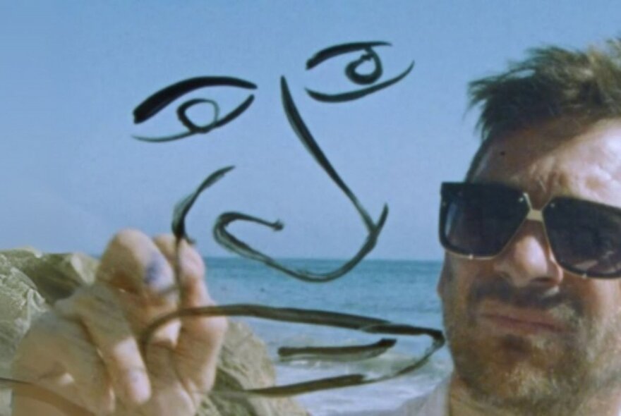 A man wearing dark sunglasses, drawing a face on the camera lens in black marker pen, with the ocean behind him.