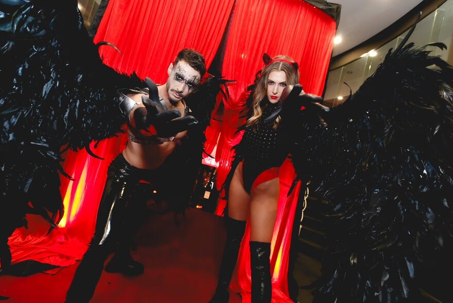 Two people dressed in elaborate Halloween inspired costumes, of black feathers, long boots, and ghostly face make up, standing in front of a red curtain inside a nightclub.