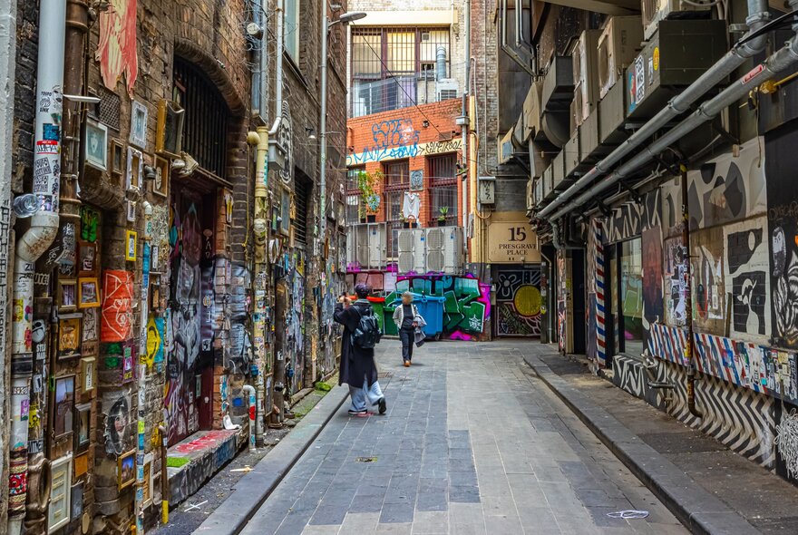 A laneway with walls covered in street art. There is a person taking photos and another person walking down the laneway.