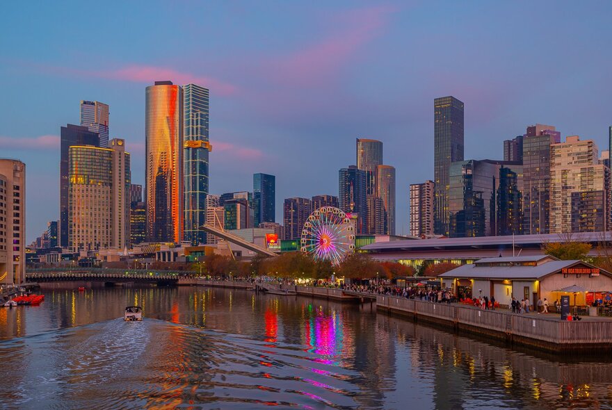 A boat travelling down the Yarra River at sunset. The city skyline and a glowing Ferris wheel are in the background.