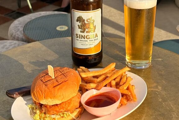 A chicken burger, fries and hot sauce on a plate, with a Singha bottle of beer and glass of beer.