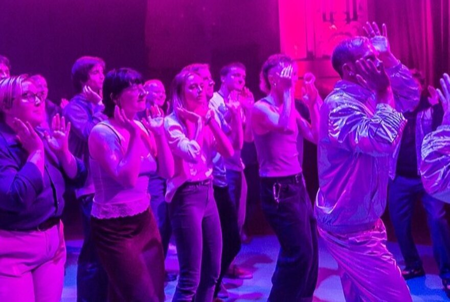 A group of people dancing in sync with their hands around their faces, lit up by a pink neon light.