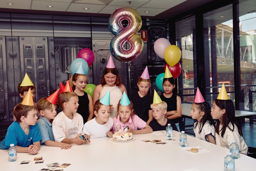 A group of children gathered around a table with a cake to celebrate an eighth birthday, inside a large room.