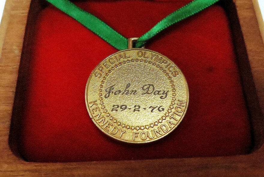 Close up of a Special Olympics gold medal with the inscription John Day and a date that reads 29-2-76, resting against a red velvet backing.
