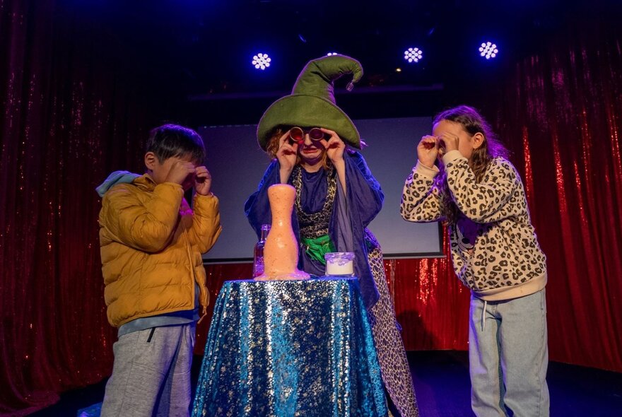 Winni Wizard, a kids comedian, performing on stage and interacting with two children standing either side of her.