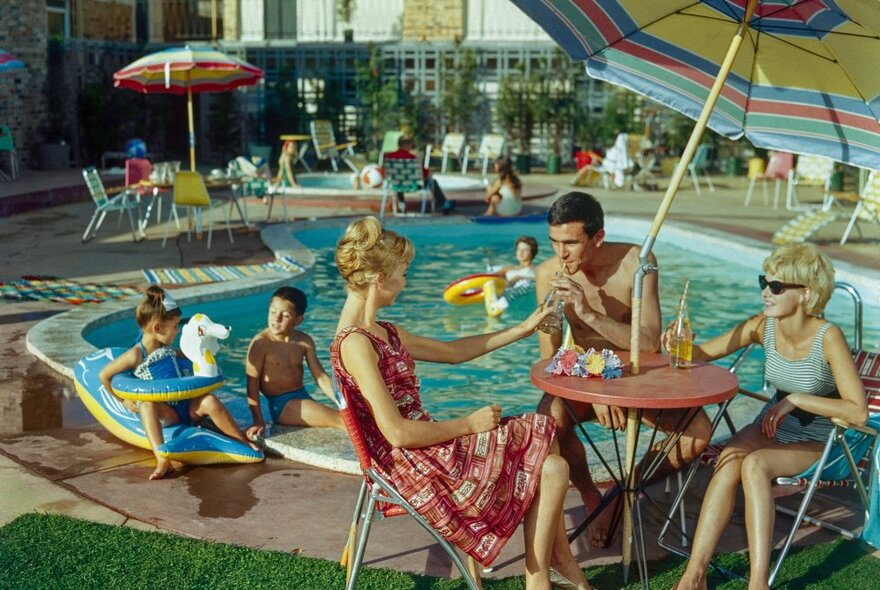 People relaxing around a motel swimming pool, sitting in deck chairs under umbrellas sipping drinks, children playing; image from the mid-1960s.