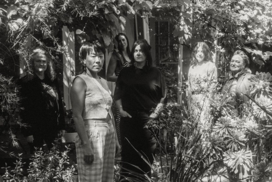 A moody black and white image of six women arranged in a garden setting in dappled light. 