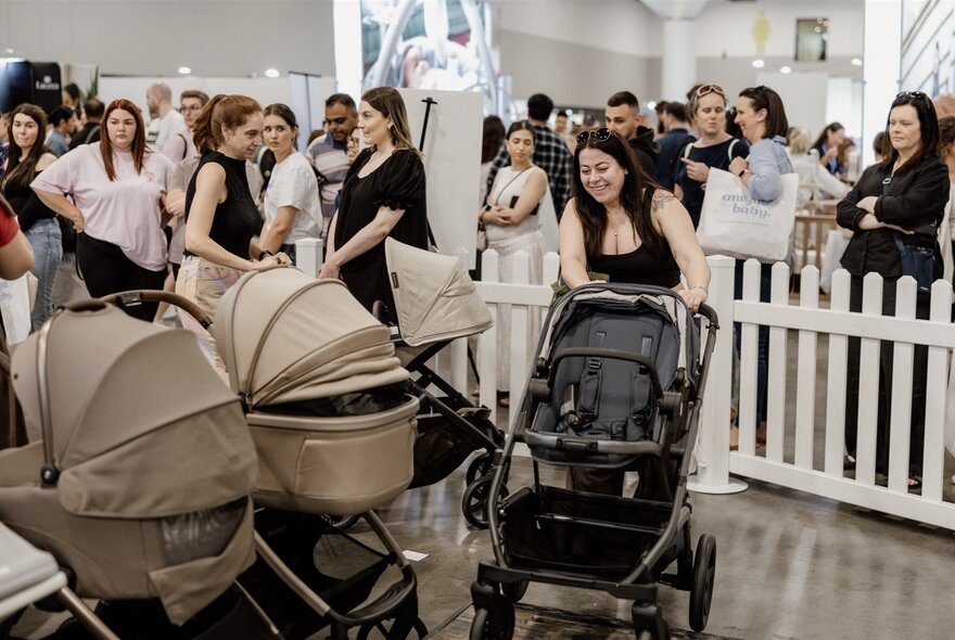 Women trying out prams and baby buggies within a small white picket fence at an expo. 