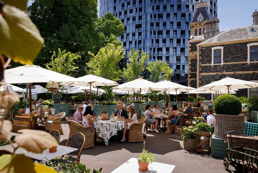 Outdoor courtyard with people sitting at tables under shade umbrellas, large shady trees with a bluestone building visible in the background.