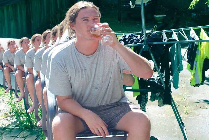 Man with his long hair tied back in a ponytail, sitting on a chair in front of a clothesline of laundry, and drinking from a glass of wine. 