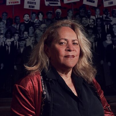 Professor Julie Andrews, an Aboriginal Australian historian, academic, and author, posed wearing a burgundy jacket and black top in front of a photo wall mural.