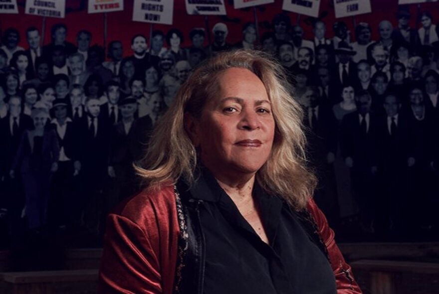 Professor Julie Andrews, an Aboriginal Australian historian, academic, and author, posed wearing a burgundy jacket and black top in front of a photo wall mural.