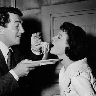 Black and white vintage photo of a man holding out a plate of spaghetti and putting a forkful into a woman's open mouth.