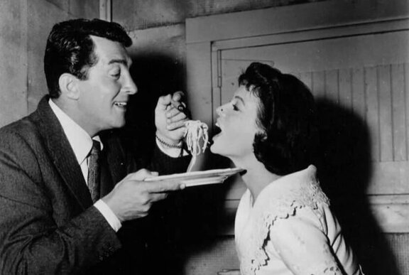 Black and white vintage photo of a man holding out a plate of spaghetti and putting a forkful into a woman's open mouth.