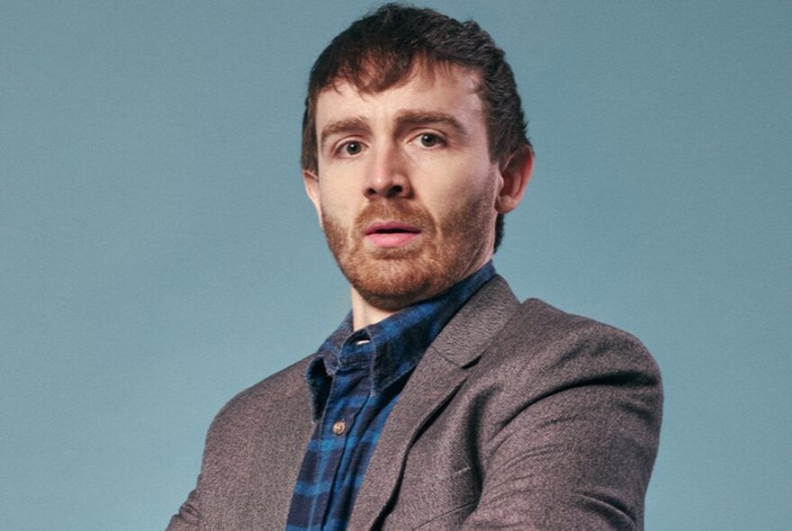 Comedian Ian Smith, wearing a blue checked shirt and a grey suit jacket, with a short beard, standing in front of a pale blue background.