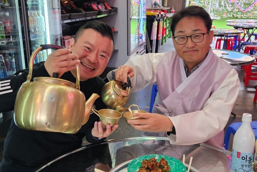 Two men smiling and pouring tea from a brass pot into small brass cups.