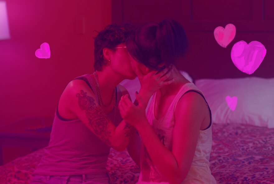 A still from the movie Egghead & Twinkie  showing two people kissing in a dark pink lit bedroom with love hearts around their heads.