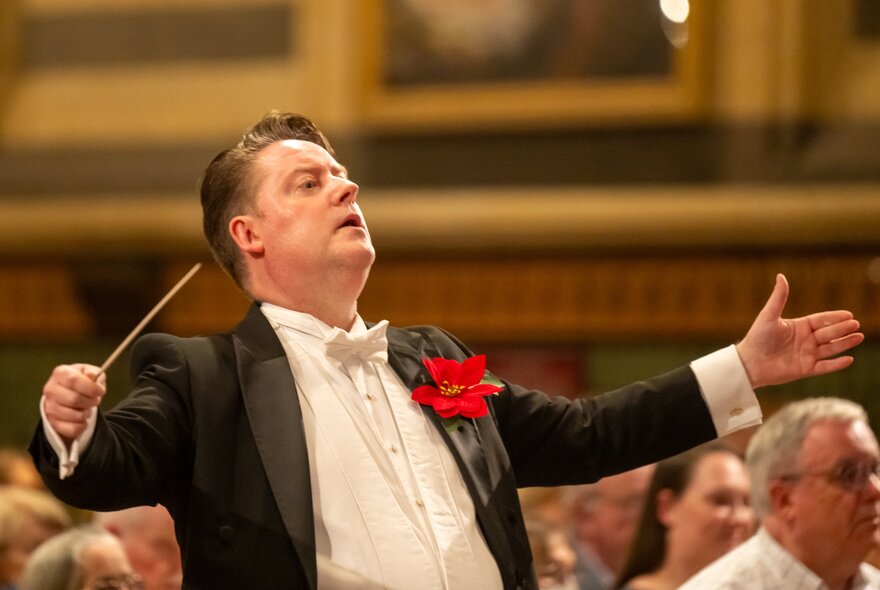 A conductor wearing a tuxedo with a red flower in his lapel, conducting with a baton.