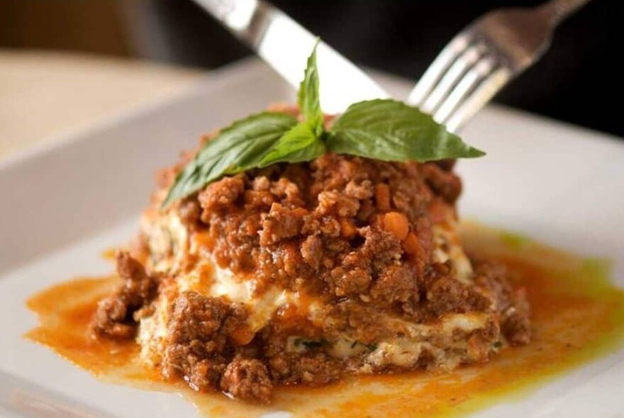 Knife and fork cutting into a meat dish garnished with basil.