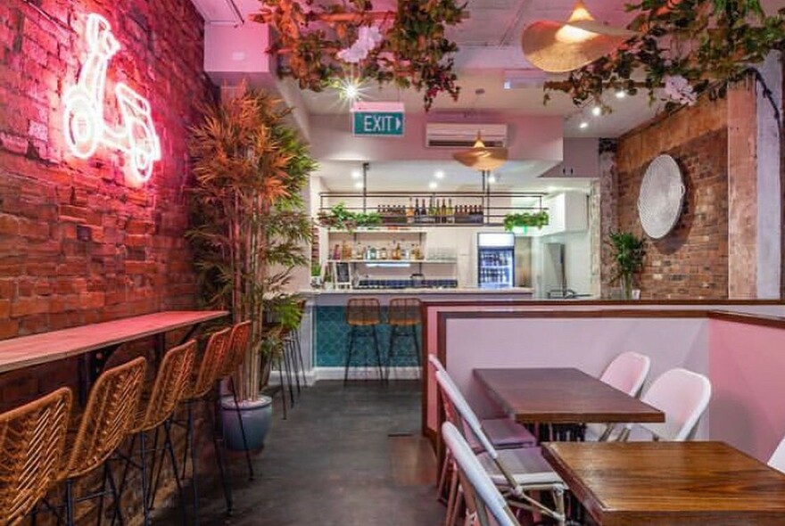 Restaurant interior with red neon motorcycle sign on a brick wall, bench and table seating, bar in the background.