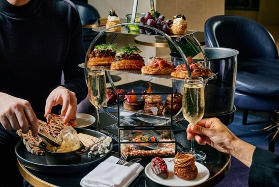 Two people enjoying High Tea from a circular tiered serving platter on a round black table with two champagne glasses.