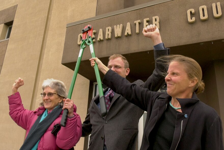 Three people outside a courtroom holding up a pair of boltcutters in celebration.